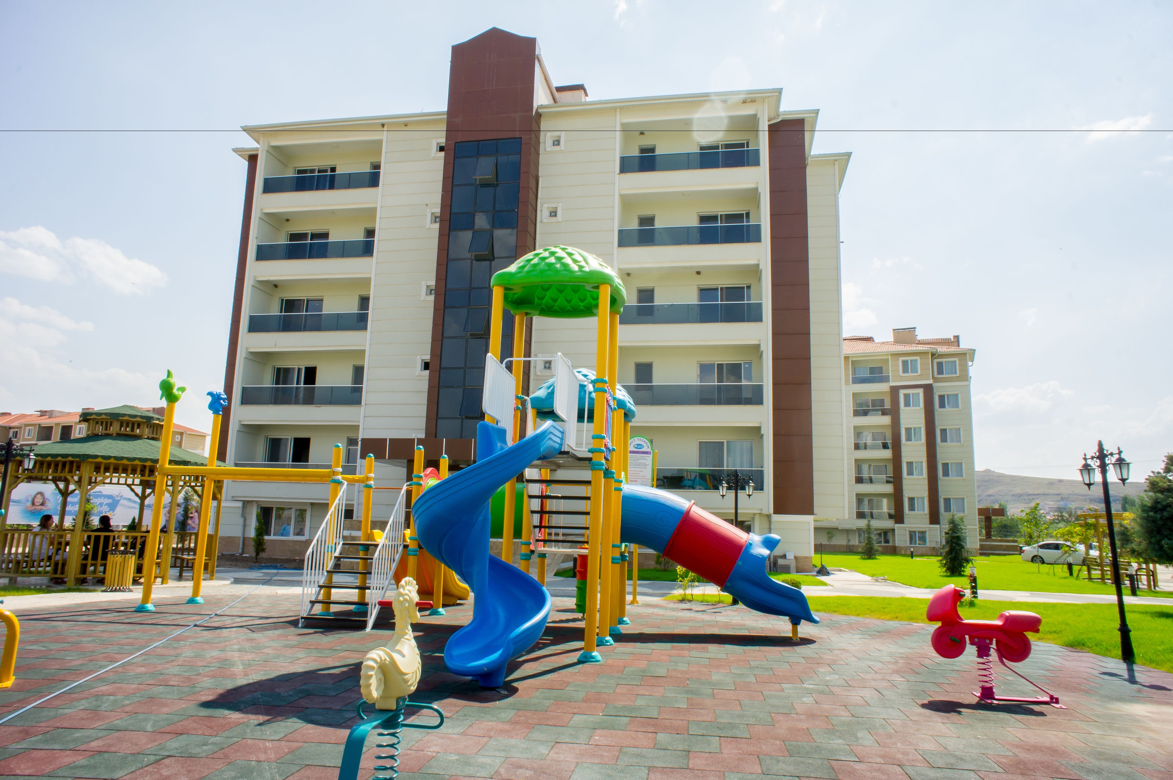 Un parque infantil con toboganes y columpios, un carrusel amarillo y rojo, y un edificio con varios balcones al fondo.