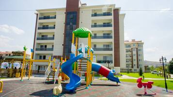 Un parque infantil con toboganes y columpios, un carrusel amarillo y rojo, y un edificio con varios balcones al fondo.