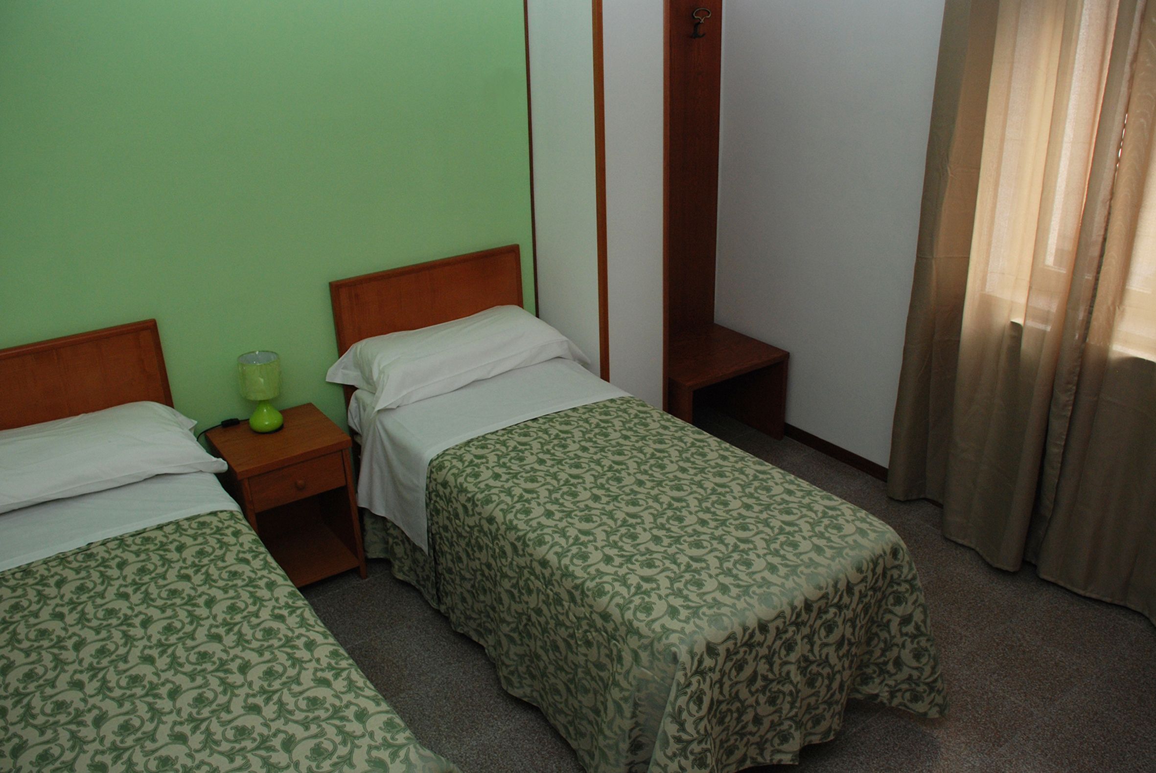 Una camera d'albergo con due letti singoli, un comodino con una lampada e un armadio in legno.
