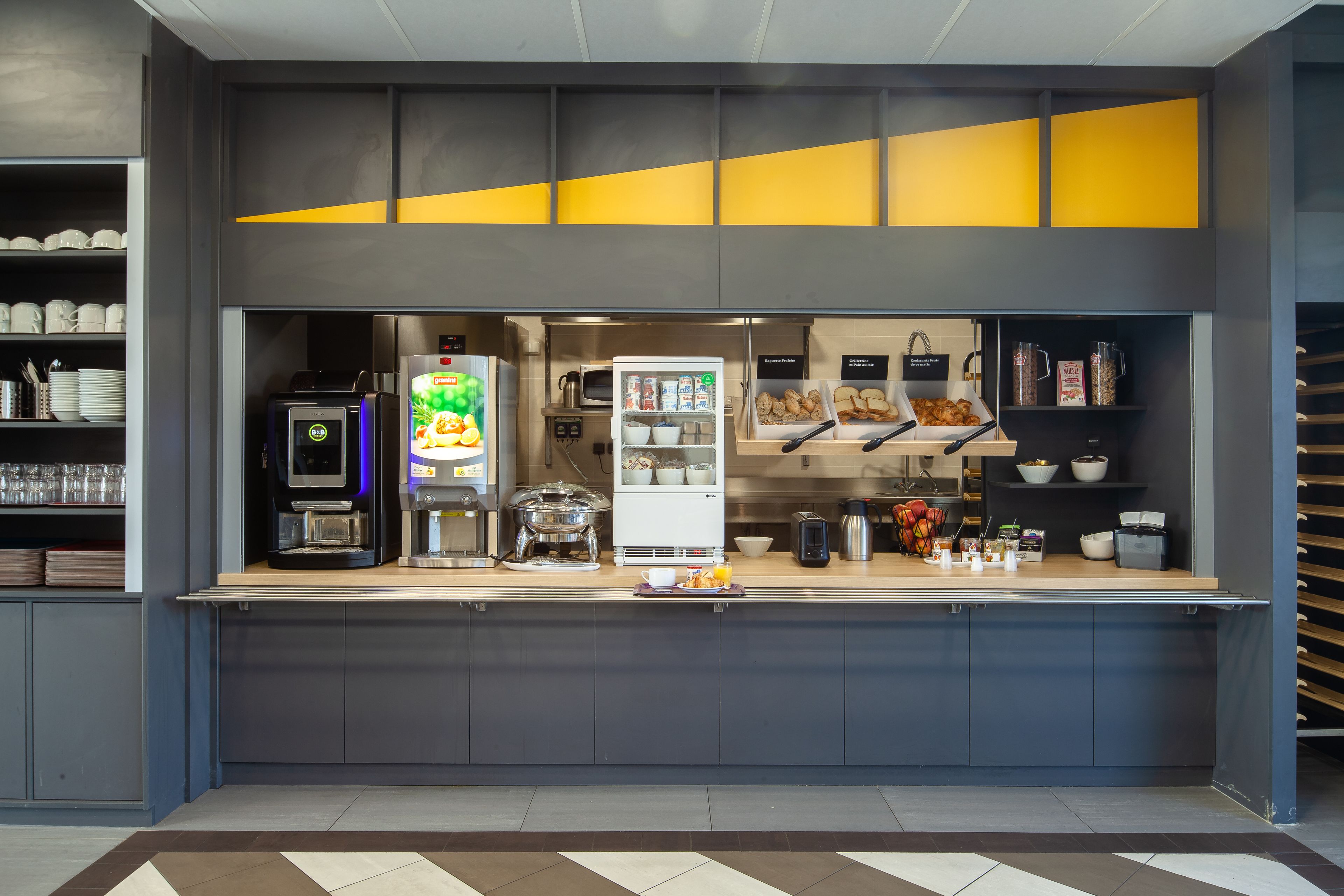 Una barra de desayuno moderna con máquinas de café, un refrigerador y diversas opciones de alimentos.