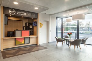 Un área de recepción de hotel con diseño moderno, que incluye un mostrador de recepción, una pantalla digital y un espacio de descanso con una mesa y sillas.