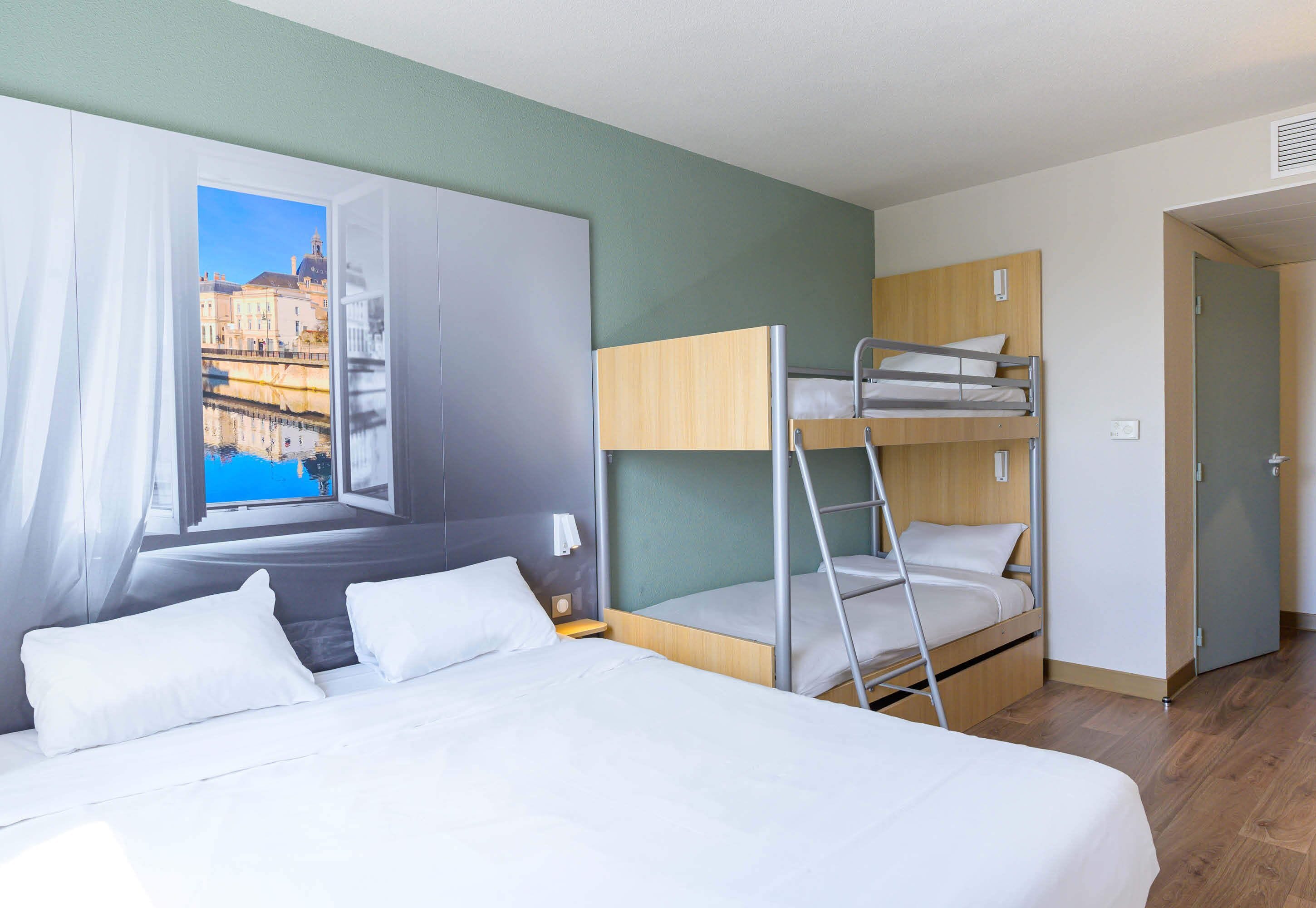 Hotellihuone, jossa on suuri sänky, kerrossänky, ikkuna, josta on näkymä rakennuksiin, ja ovi.