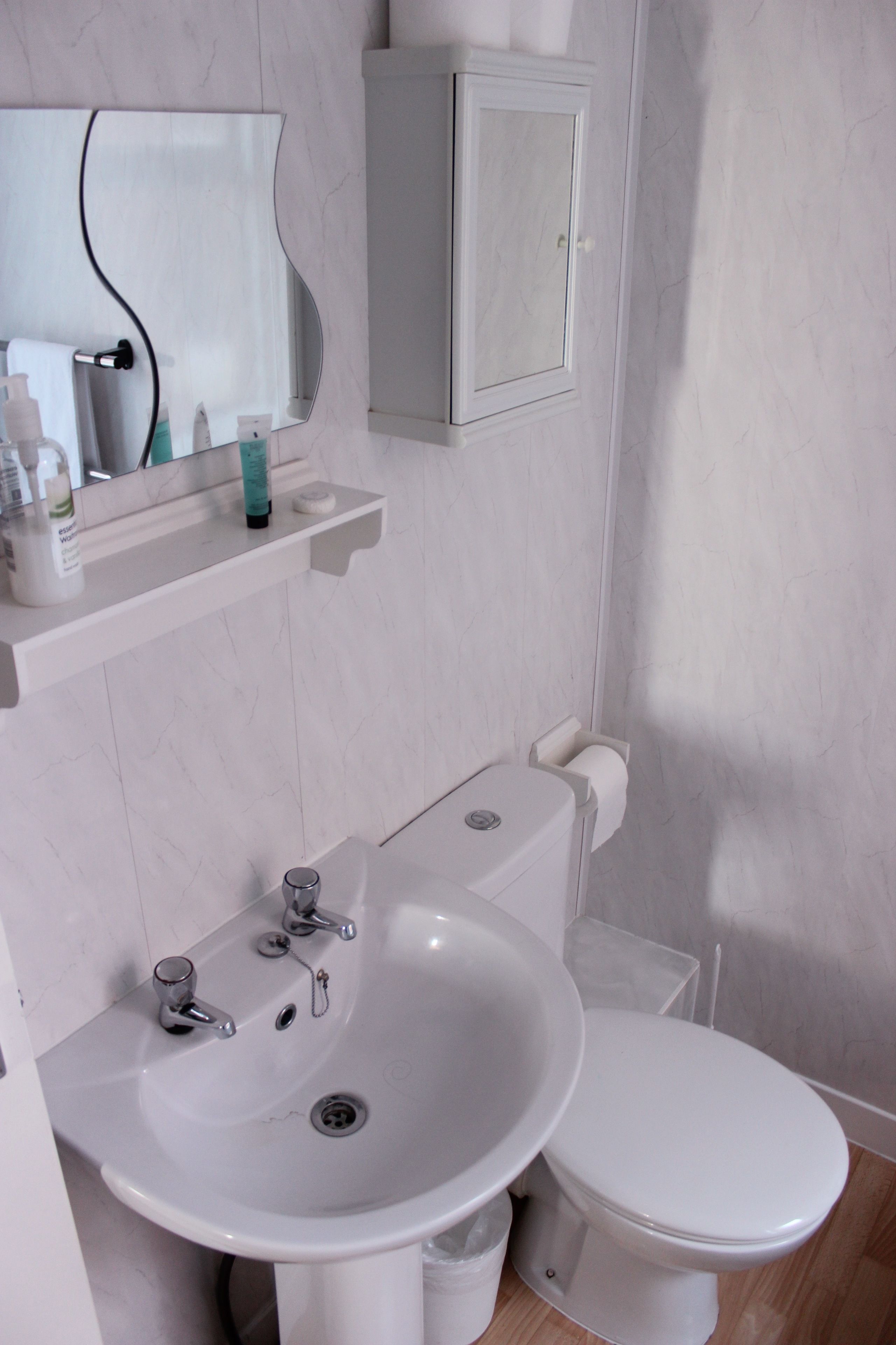 A bathroom with a white sink, toilet, and a mirror above the sink. There is a small cabinet above the toilet and a shelf with toiletries.