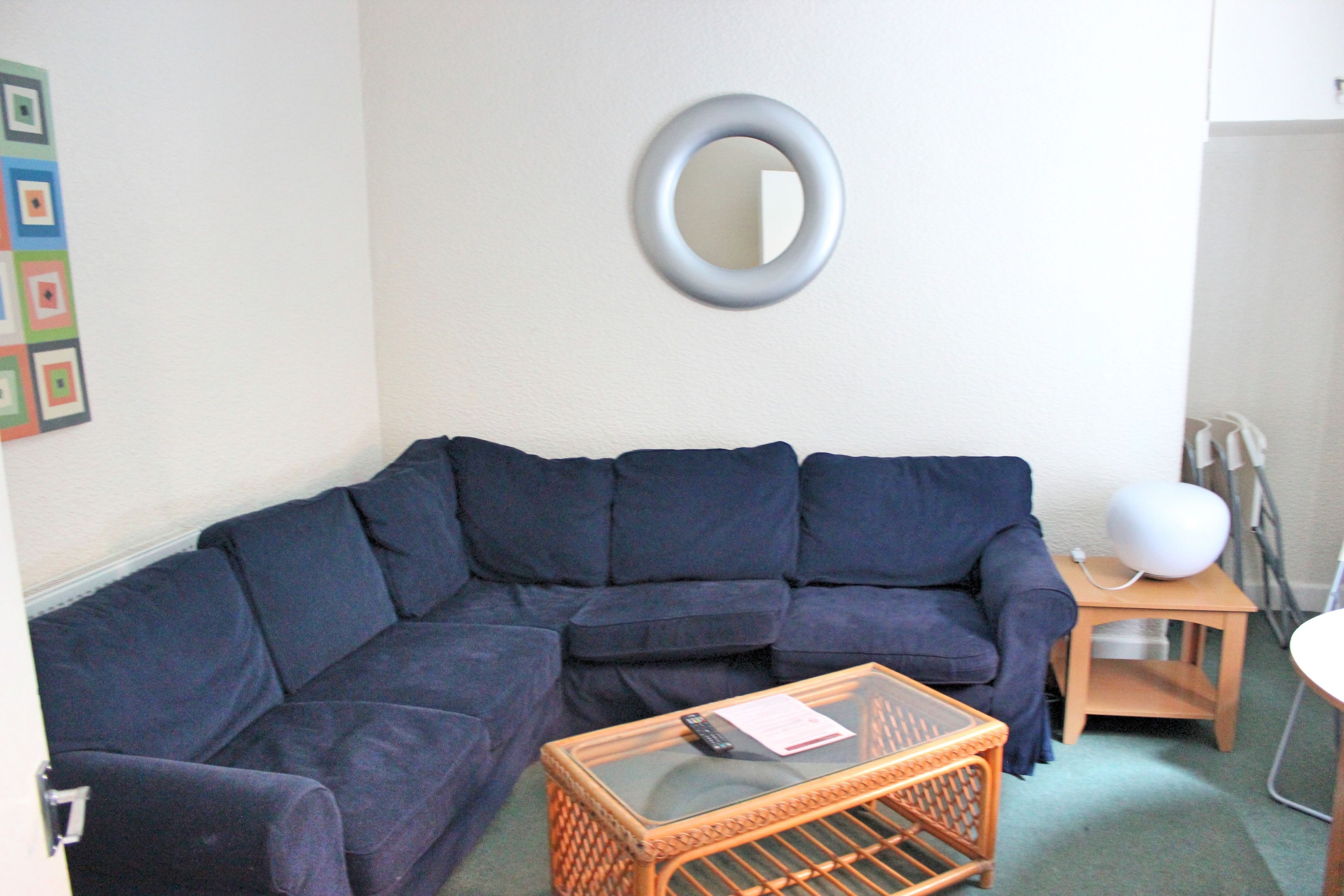 A living room with a dark blue sectional sofa, a glass-top coffee table, a round mirror on the wall, and a small wooden side table with a lamp.