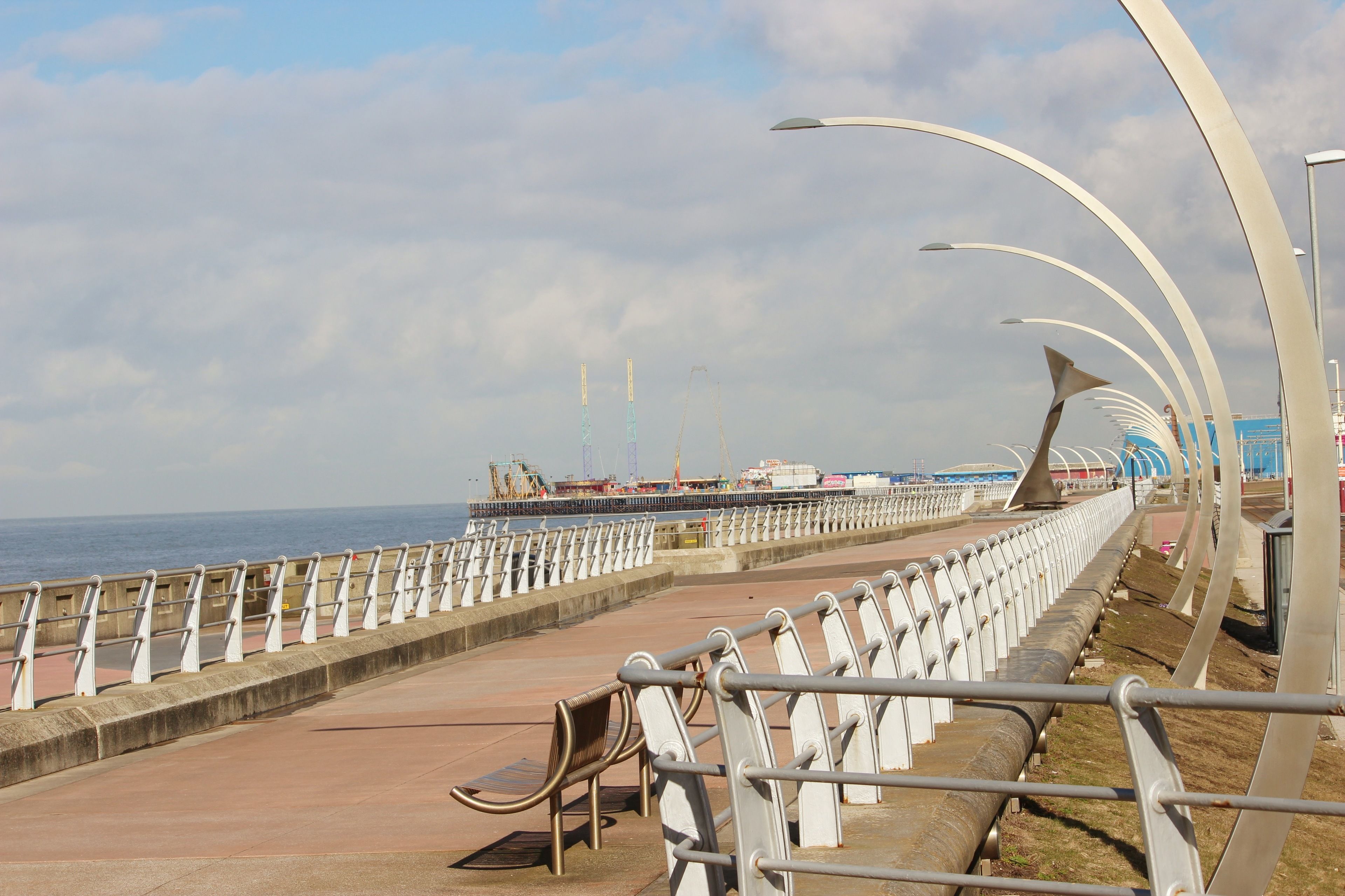 Eine Küstenpromenade mit Metallgeländer, Bänken und Blick auf das Meer sowie entfernte Gebäude.
