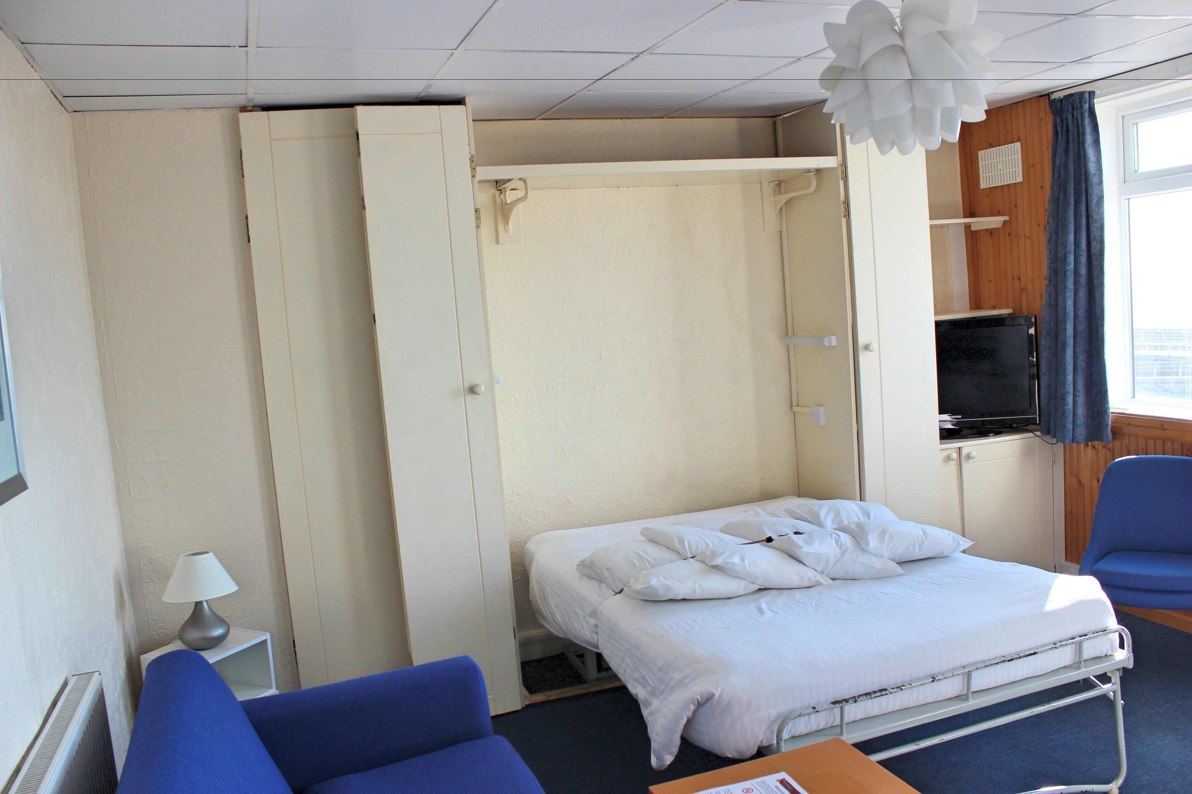 Ein Einzelzimmer mit einem Bett, einem Sessel, einem Fernseher und einer Lampe.