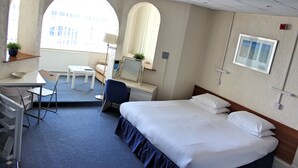 A hotel room with a bed, desk, chair, and a window with a view of buildings.