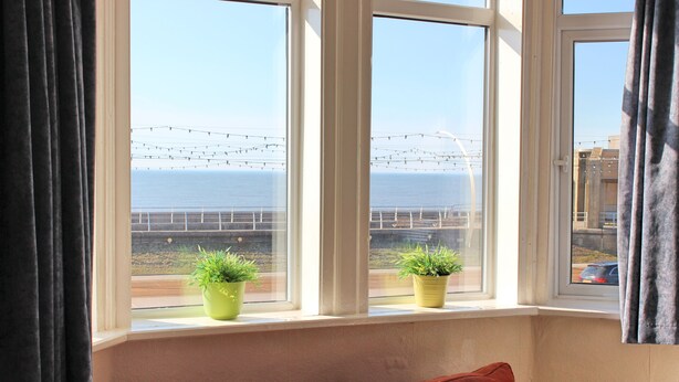 A room with a view of the sea, two potted plants on the windowsill, and a red cushion on the sofa.