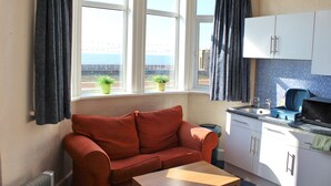A room with a red sofa, a wooden table, a kitchen with white cabinets, and a view of the sea through large windows.