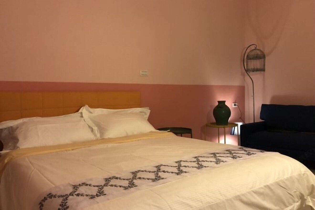 A hotel room with a bed, a small table with a green vase, a floor lamp, and a dark-colored sofa.