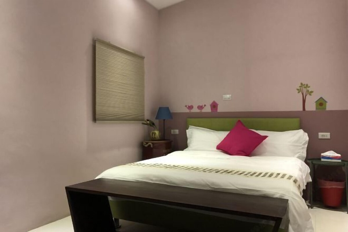 A bedroom with a bed, a nightstand, a lamp, a trash bin, and a window with blinds.