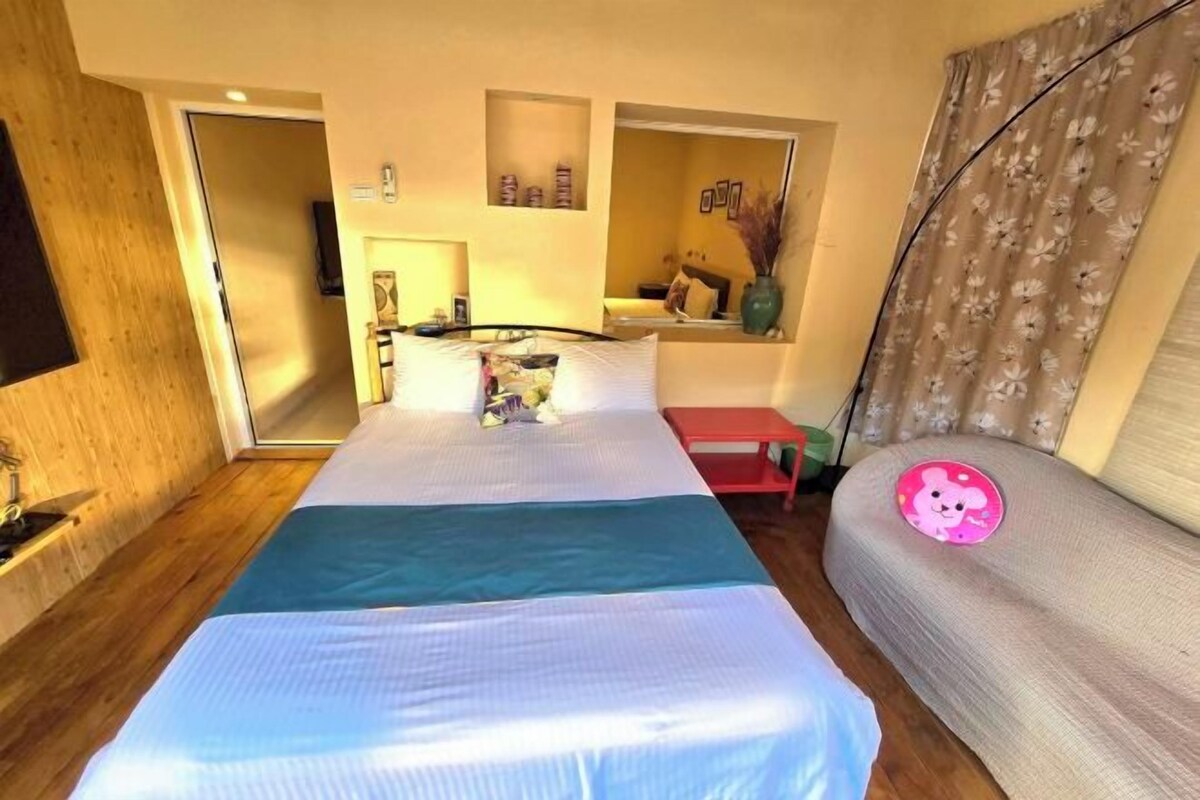 Un dormitorio con cama, una mesita de noche roja, un espejo y una ventana con cortinas.