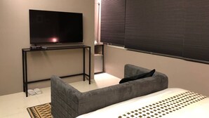 A modern hotel room with a bed, a gray sofa, a TV on a stand, and a window with blinds.