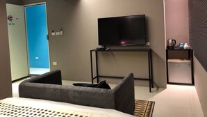 A modern hotel room with a bed, a TV on a stand, a small table, and a kitchenette area.