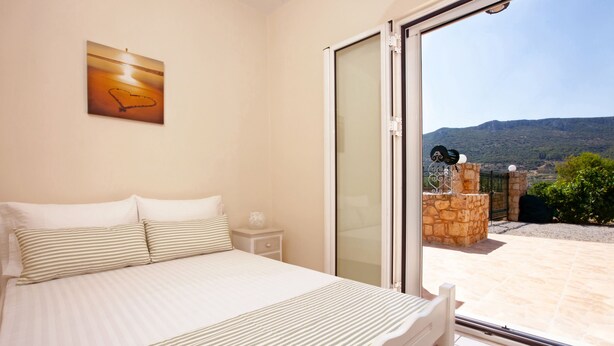 A bedroom with a bed, a nightstand, and a view of a mountainous landscape through a sliding glass door. - Laconia Domus - Papadianika (Monemvasia)