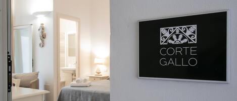 Habitación de hotel con cama, escritorio y baño visible a través de una puerta abierta. Un cartel en la pared dice 'Corte Gallo'.