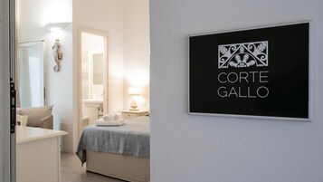 Habitación de hotel con cama, escritorio y baño visible a través de una puerta abierta. Un cartel en la pared dice 'Corte Gallo'.