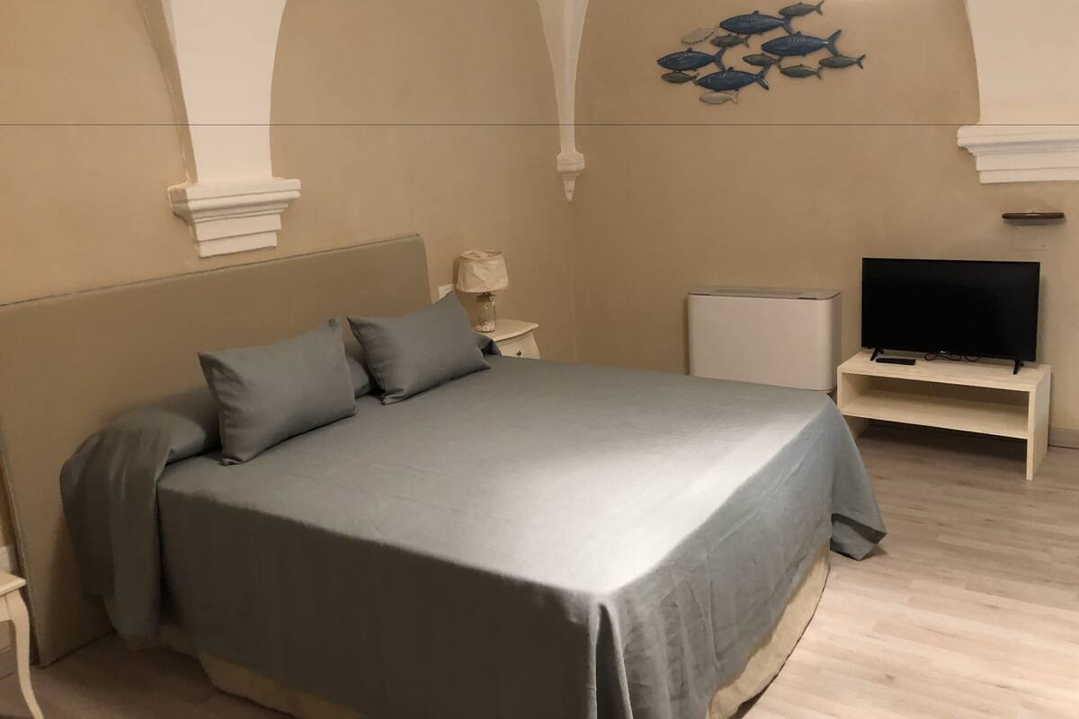 Un dormitorio con cama, televisor sobre un mueble bajo, una mesita de noche con lámpara y un adorno de peces en la pared.