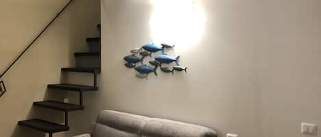 A living room with a grey sofa, a small round table with a lantern, a wall-mounted fish decoration, a staircase, and a ceiling light.