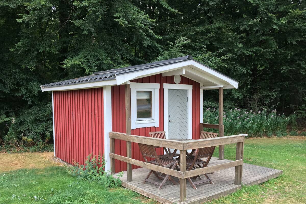 A small red wooden cabin with a porch and two doors, surrounded by greenery.