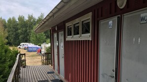 A wooden deck with a metal railing, a red building with WC signs, and a tent in the background. - Torne Camping (Lönashult)