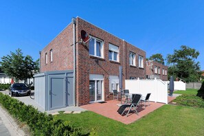 Ein zweistöckiges Backsteinhaus mit einer Terrasse, die einen Tisch und Stühle beinhaltet, einem geparkten Auto davor und einem kleinen Schuppen.