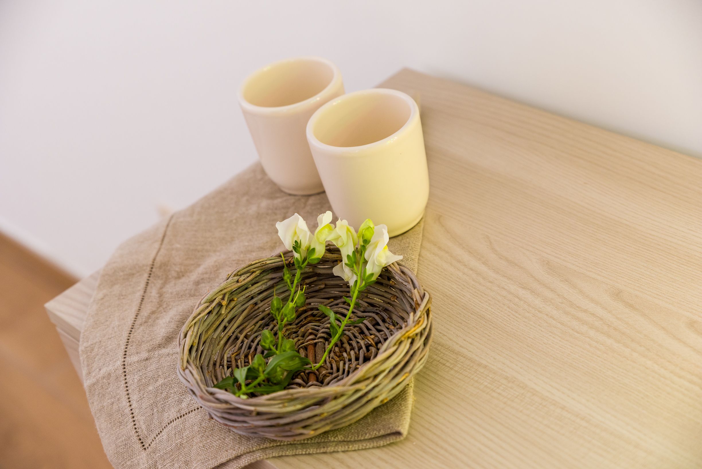 A wicker basket with flowers on a beige cloth, two plain cups, and a wooden surface.