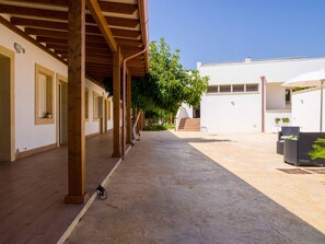 A covered outdoor area with wooden beams, a concrete floor, and a white building with windows and shutters.