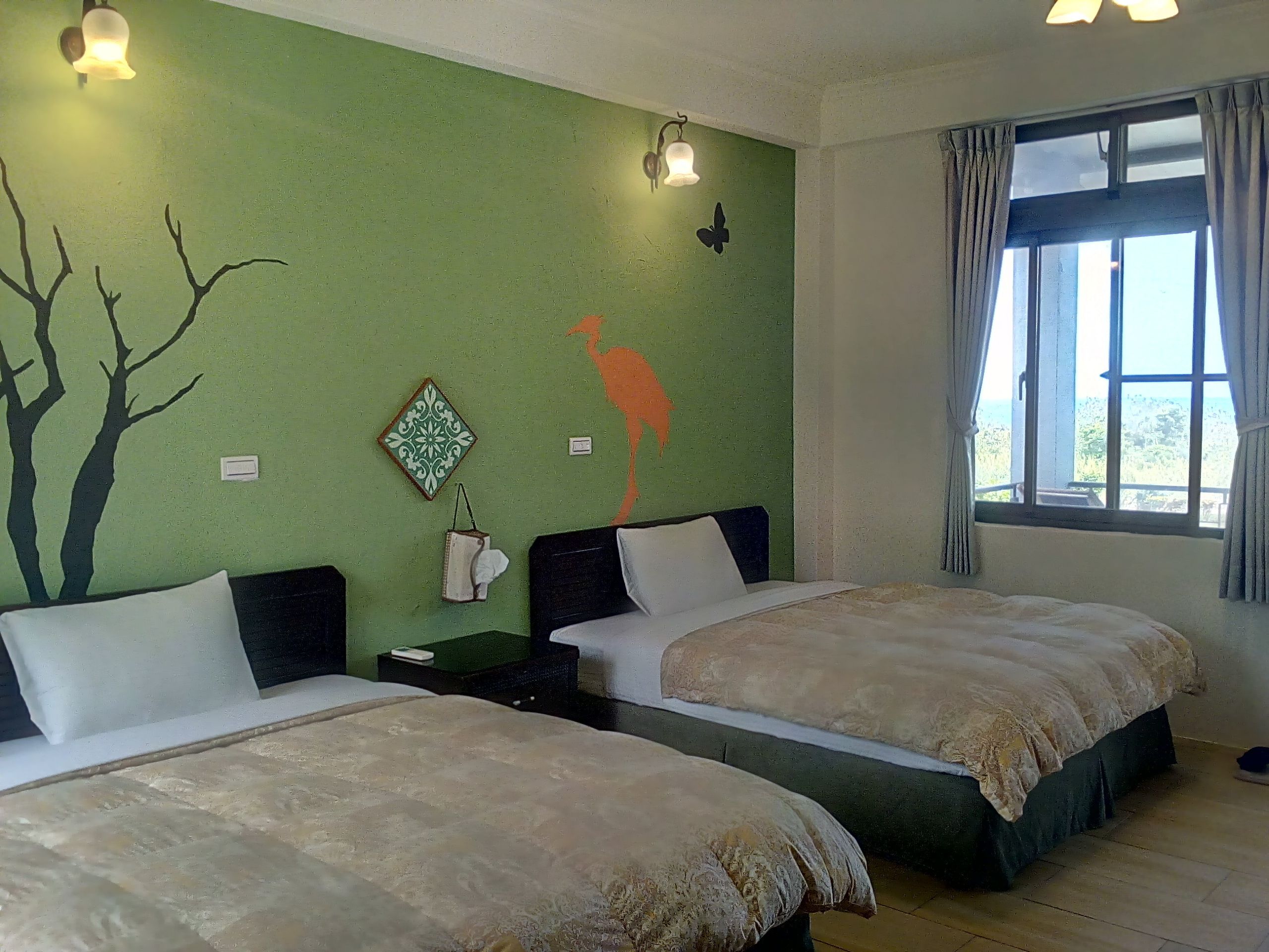 Habitación con dos camas, pared verde con un mural de árbol y mariposa, ventana con cortinas y mesita de noche con lámpara.