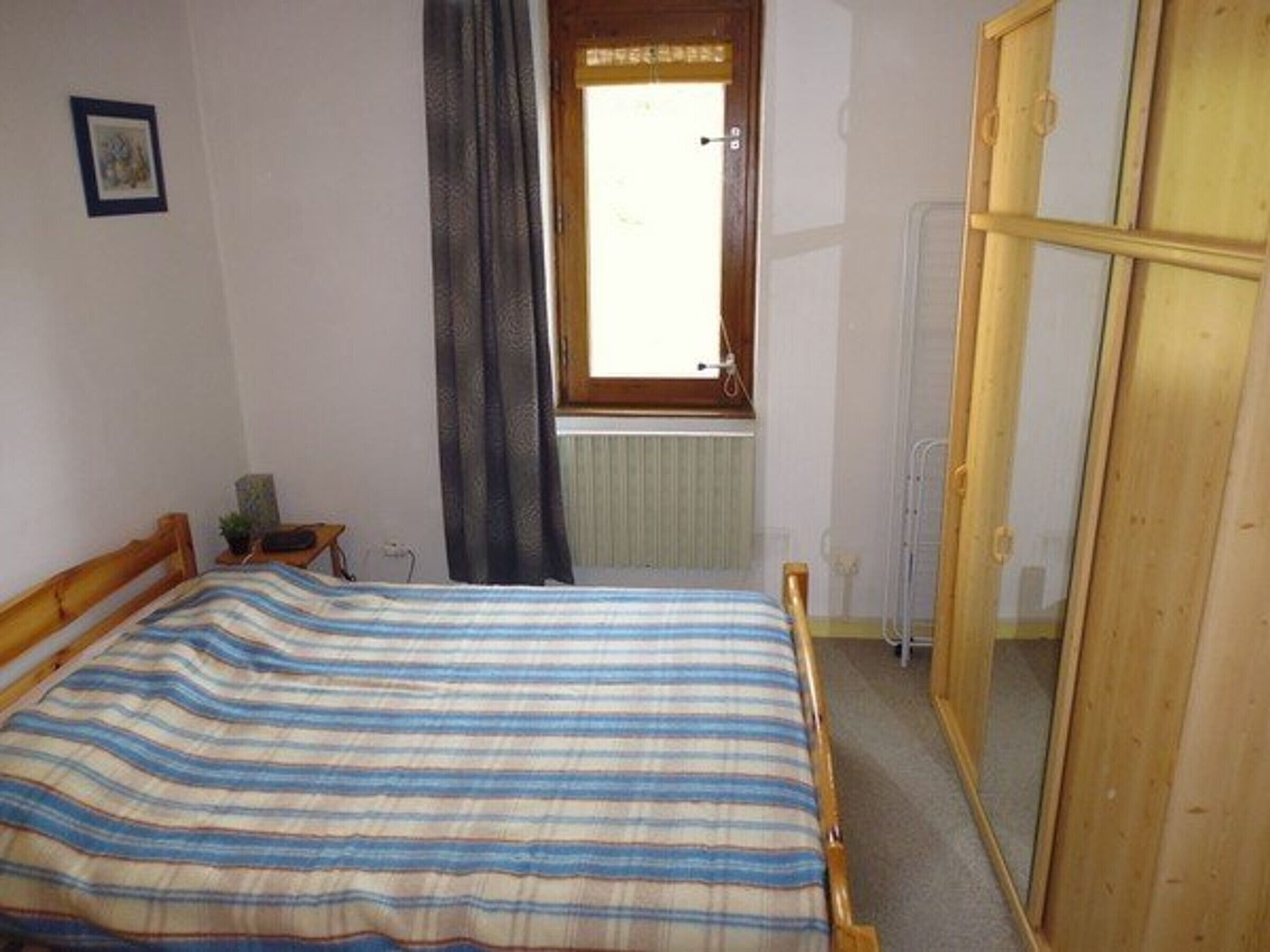 A bedroom with a bed, a window with curtains, a radiator, and a wooden wardrobe.