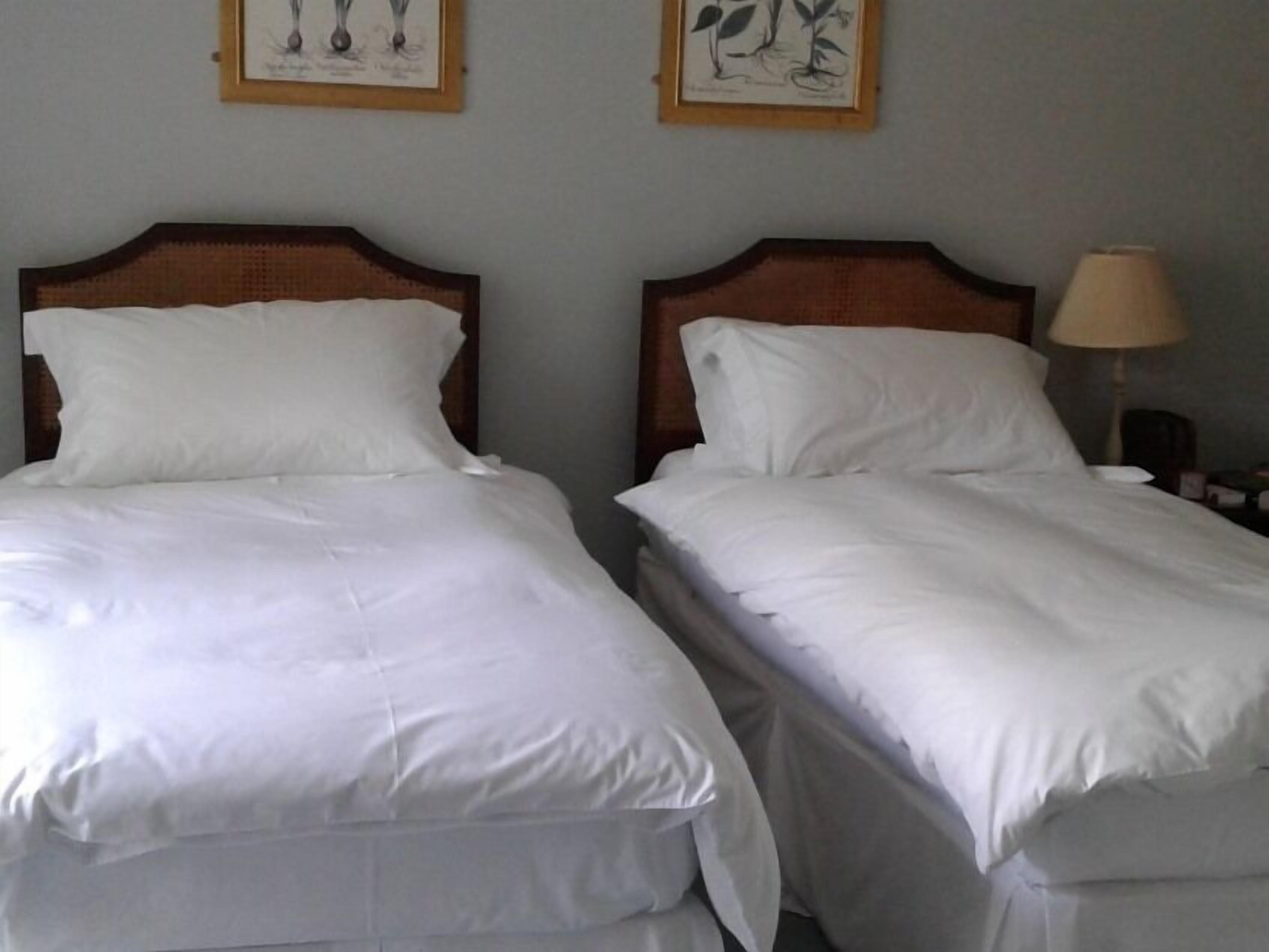 Two neatly made beds with white linens and wooden headboards, a lamp on the nightstand, and framed botanical prints on the wall.