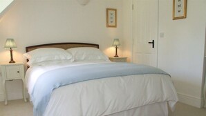 A neatly made bed with white bedding and a blue accent, two bedside tables with lamps, and a door with a handle. - Well Cottage B&B (Cirencester)