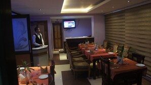 Een restaurant met meerdere tafels gedekt voor diners, een aan de muur bevestigde televisie en een ober die bij de balie staat.