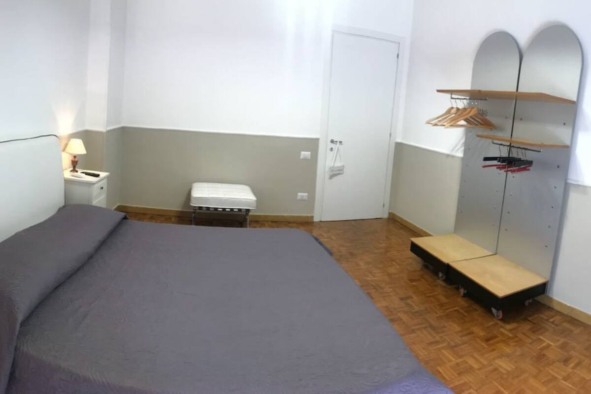 A bedroom with a bed, bedside table, lamp, bench, wardrobe, and a TV.