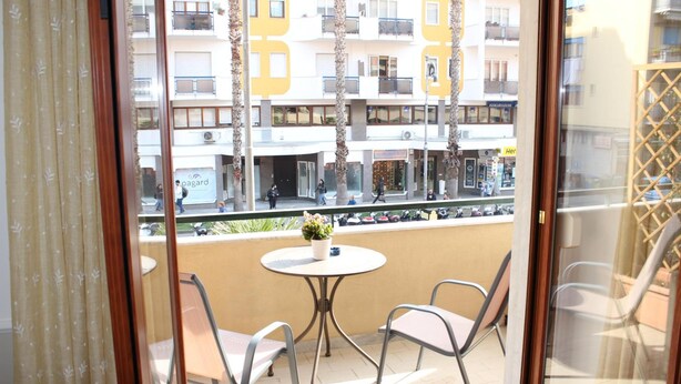 Balcón con dos sillas y una mesita, con vista a una calle con edificios y comercios.