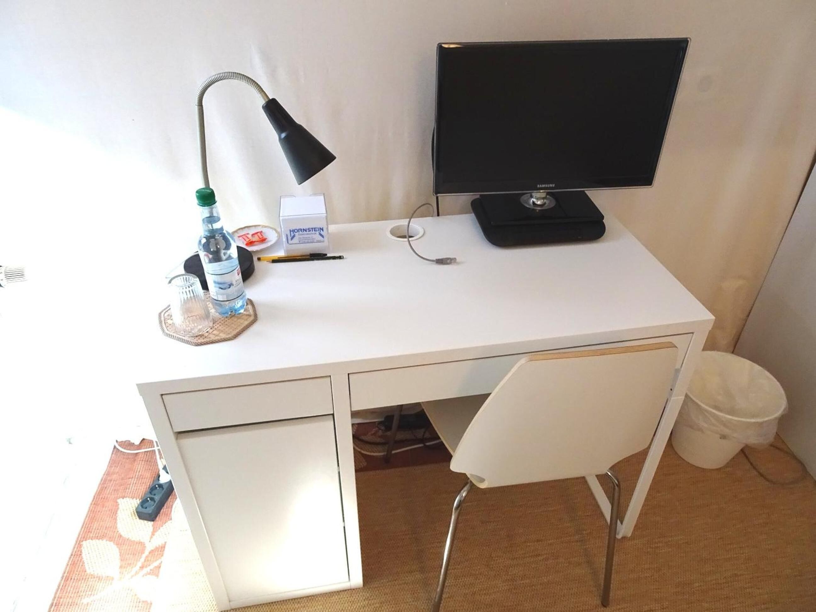 Un bureau blanc avec un écran d’ordinateur, une lampe, une bouteille d’eau, une boîte de mouchoirs et une poubelle.