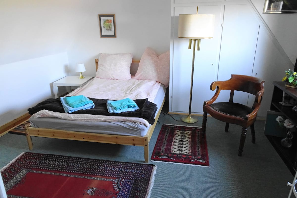 A neatly arranged bedroom with a bed, bedside table, lamp, chair, and rugs.