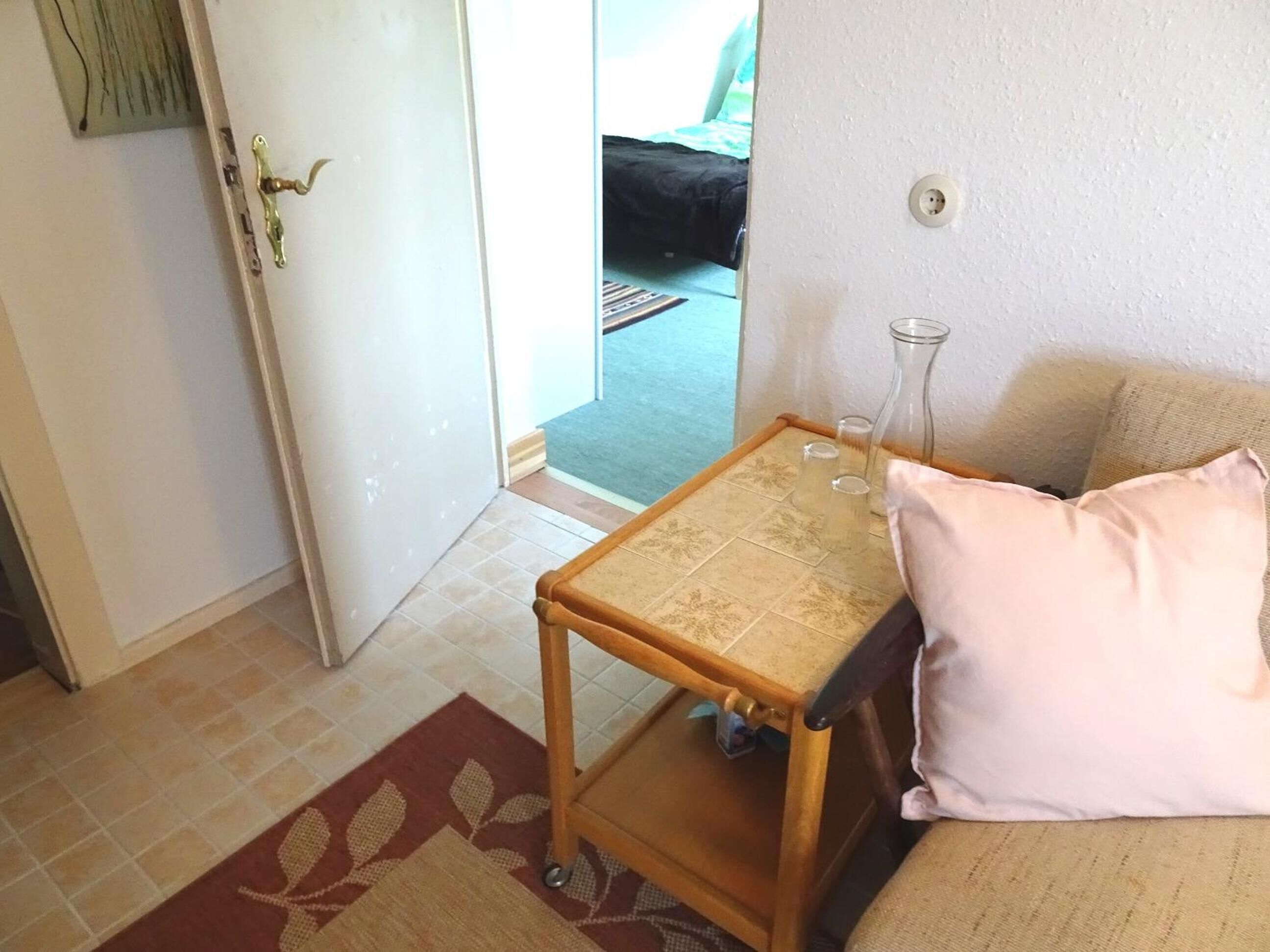 Ein kleiner Raum mit einem Holztisch, einem rosafarbenen Kissen und einer Glasvase auf einem Regal.