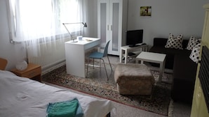 A hotel room with a bed, a desk with a computer, a television, a sofa, and a patterned rug. - Haus Fermate (Wilhelmsfeld)