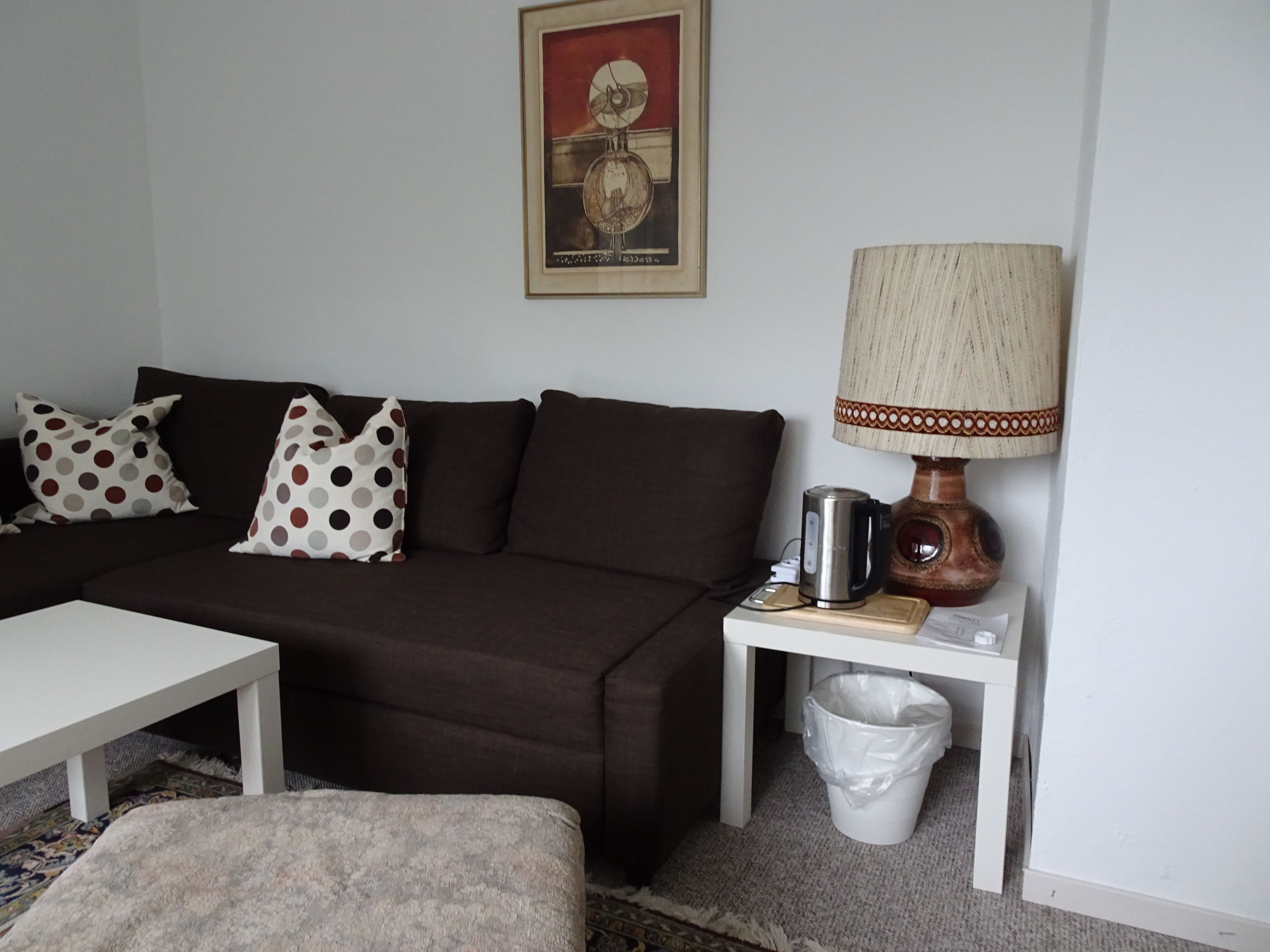 Uma sala de estar com um sofá marrom, uma mesa de centro branca e uma luminária em uma mesinha lateral.