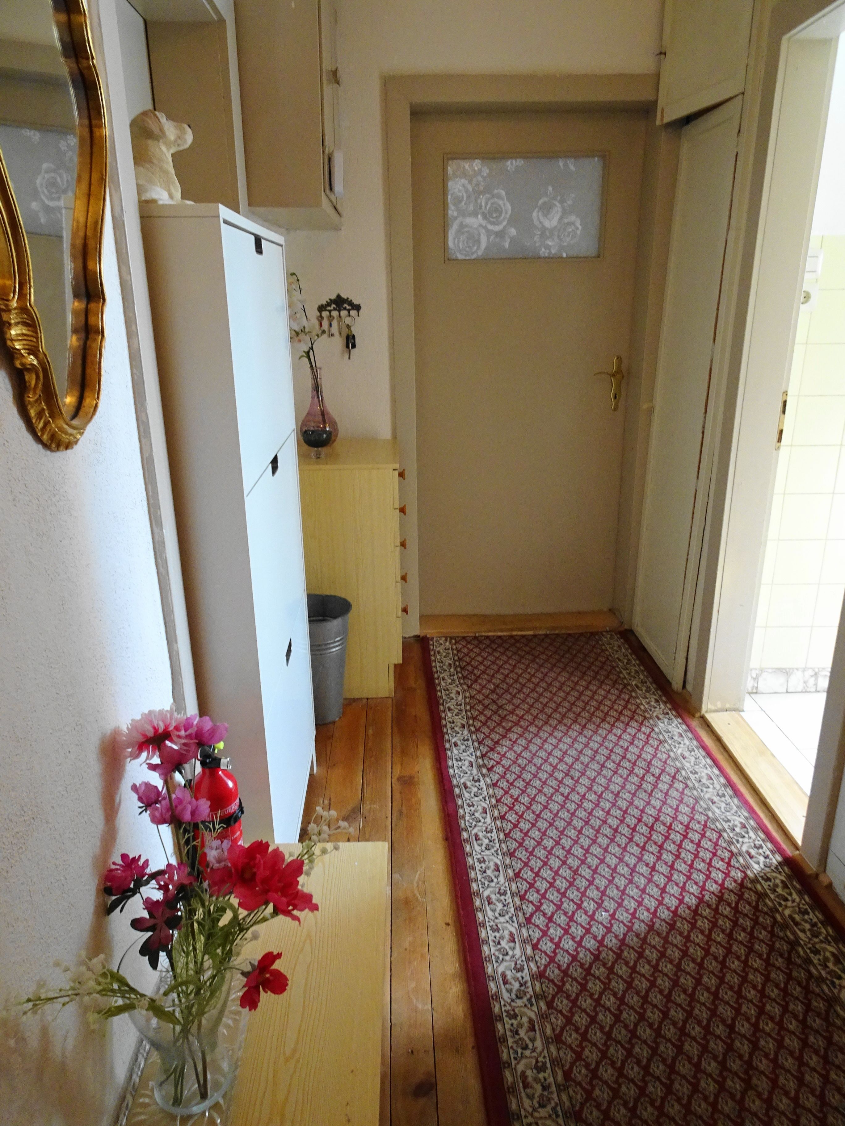 Ein Flur mit einem gemusterten Teppich, Holzboden, einem kleinen Schrank und einer Vase mit Blumen.