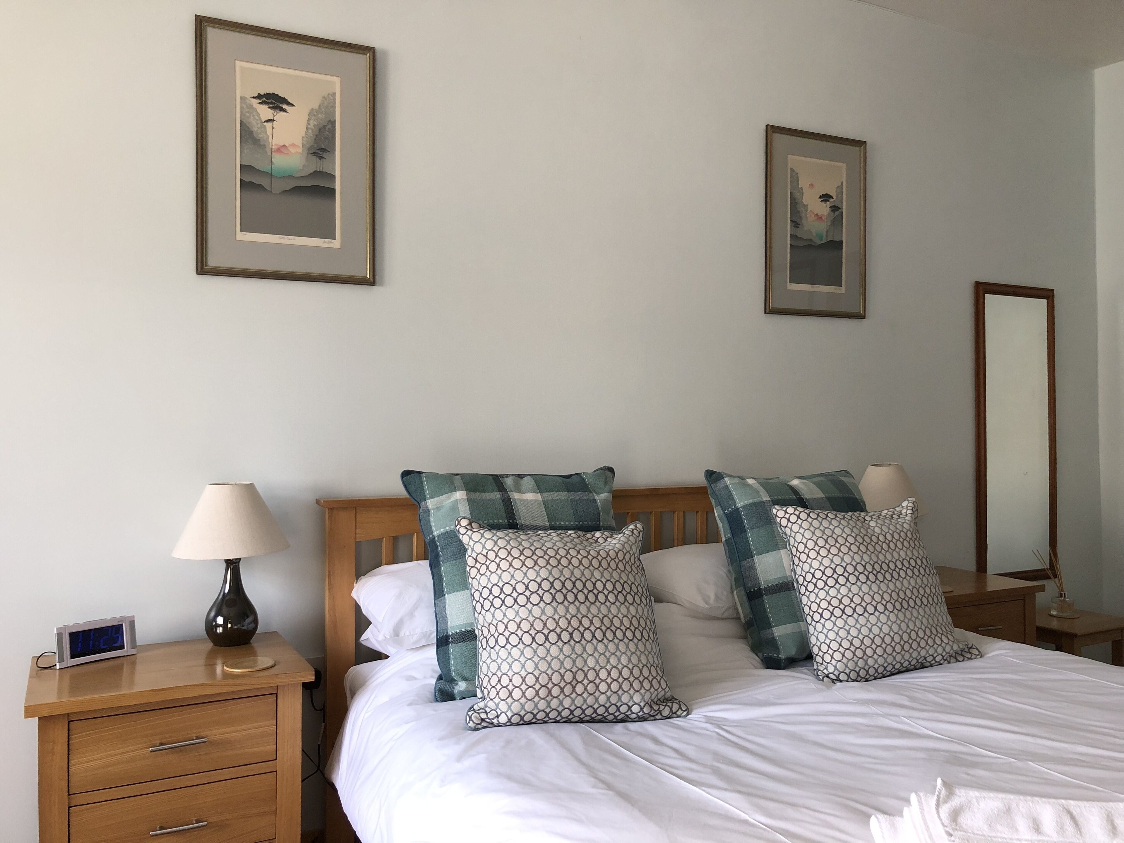A neatly arranged bedroom with a bed, two bedside tables, a nightstand, a mirror, and two framed pictures on the wall.