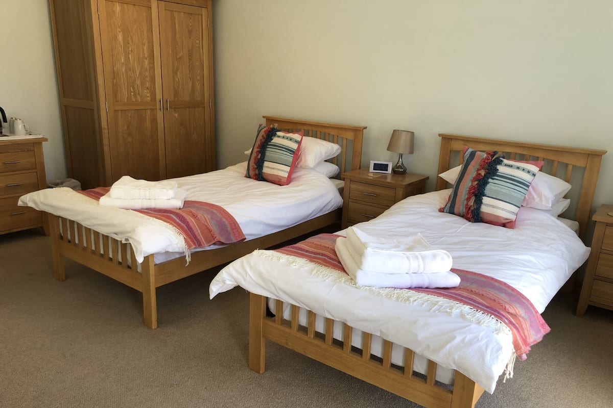Two wooden beds with white linens, striped blankets, and colorful pillows. There are wooden nightstands with lamps and a small table with a clock. A wooden wardrobe is visible in the background.