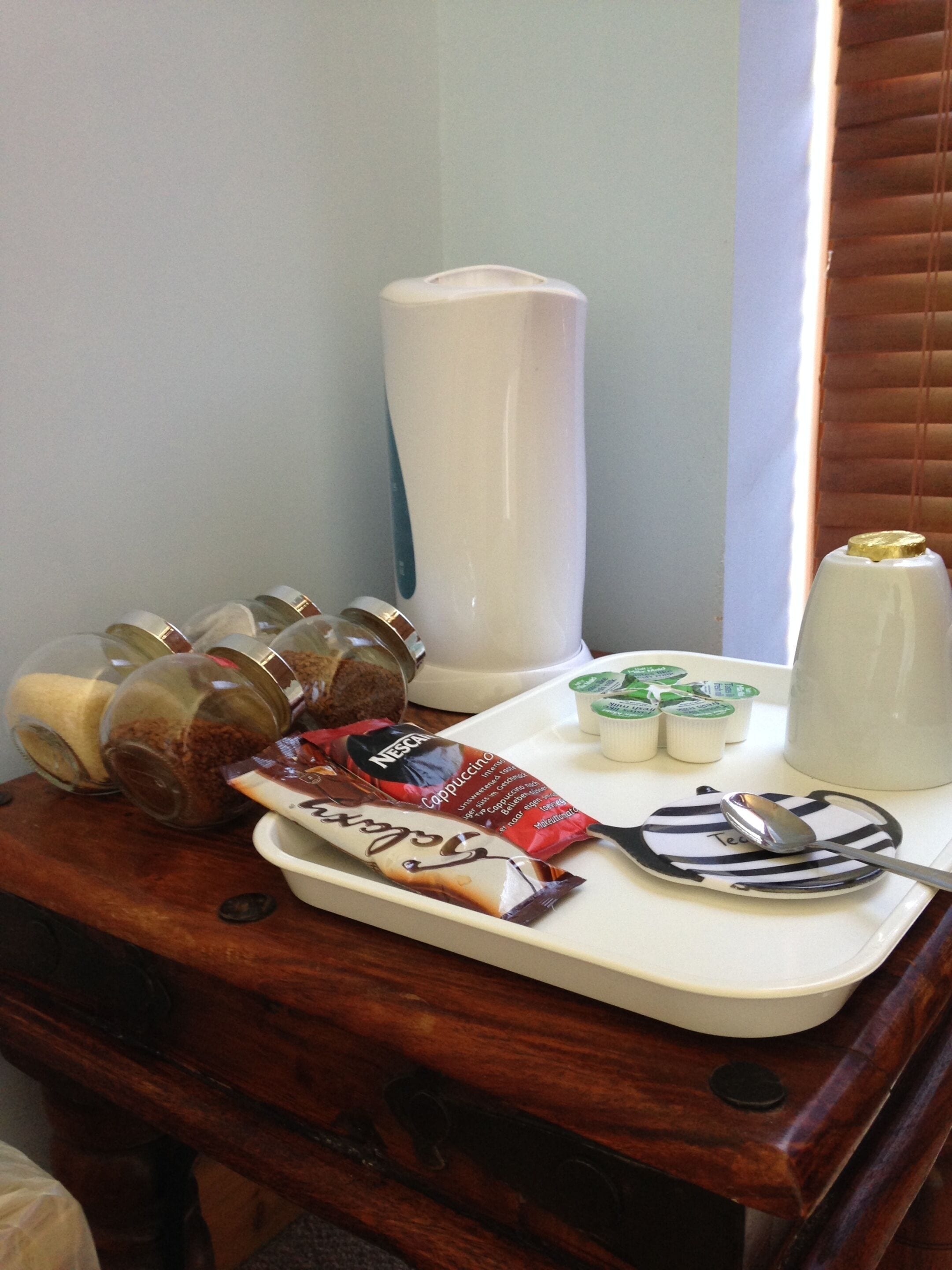 A tray with food items, including a chocolate bar, a packet of biscotti, and a container of yogurt, on a wooden table near a window with blinds.