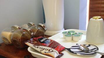 A tray with food items, including a chocolate bar, a packet of biscotti, and a container of yogurt, on a wooden table near a window with blinds.