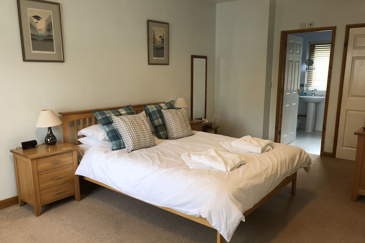A neatly made bed with white linens, a wooden headboard, and plaid-patterned pillows. There is a bedside table with a lamp and a small drawer. The room has light-colored walls and a carpeted floor.