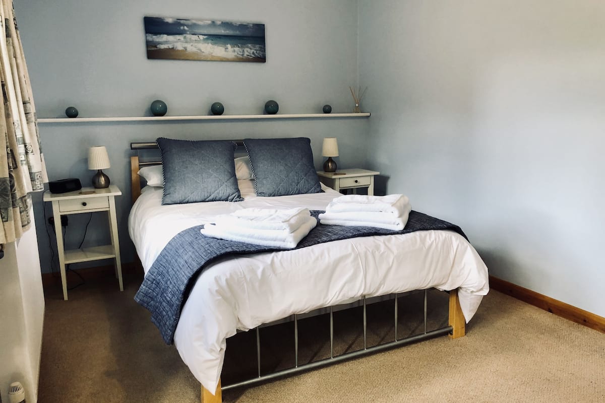 A neatly made bed with white linens and a blue blanket, flanked by two bedside tables with lamps, a wall-mounted shelf with decorative items, and a painting of a beach scene above the bed.