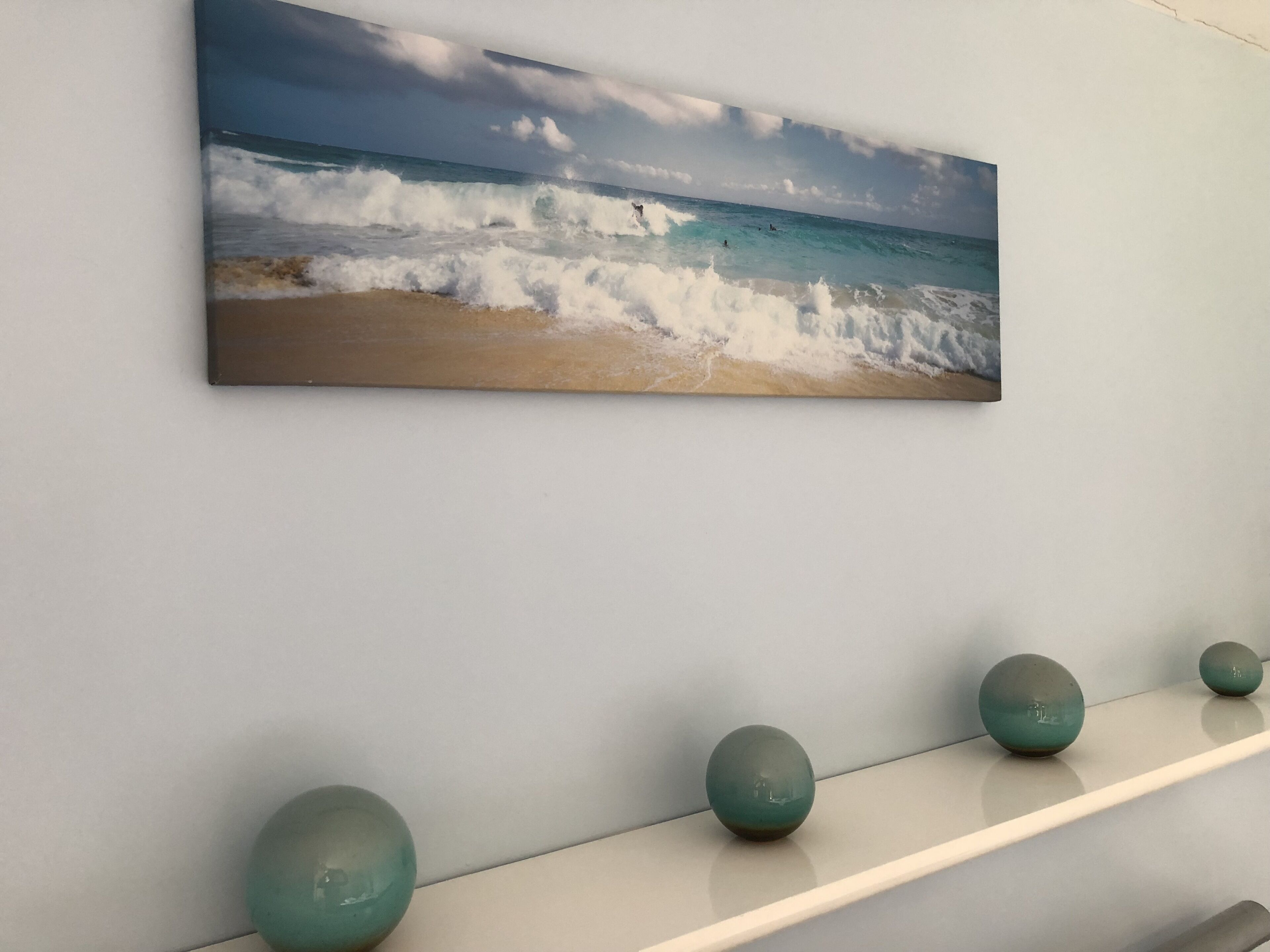 A wall-mounted painting of a beach scene with waves and a clear sky. Below the painting, there is a white shelf with three spherical objects.