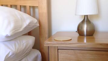Een houten bed met witte lakens, een nachtkastje met een lamp en een ladekast.