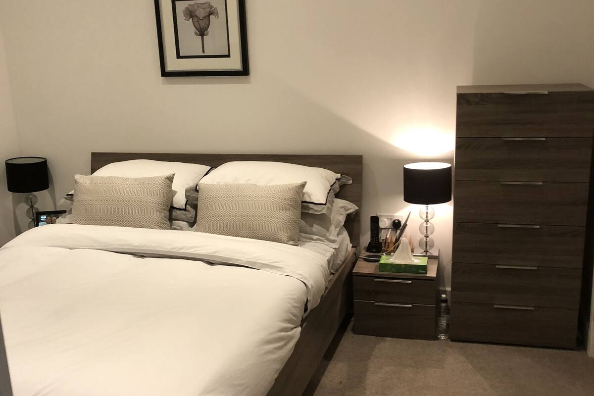 A neatly made bed with white linens, a bedside table with a lamp, a dresser, and a framed picture on the wall.
